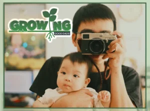 A dad holds a baby and a camera, holding the baby up in front of a mirror to snap a picture.